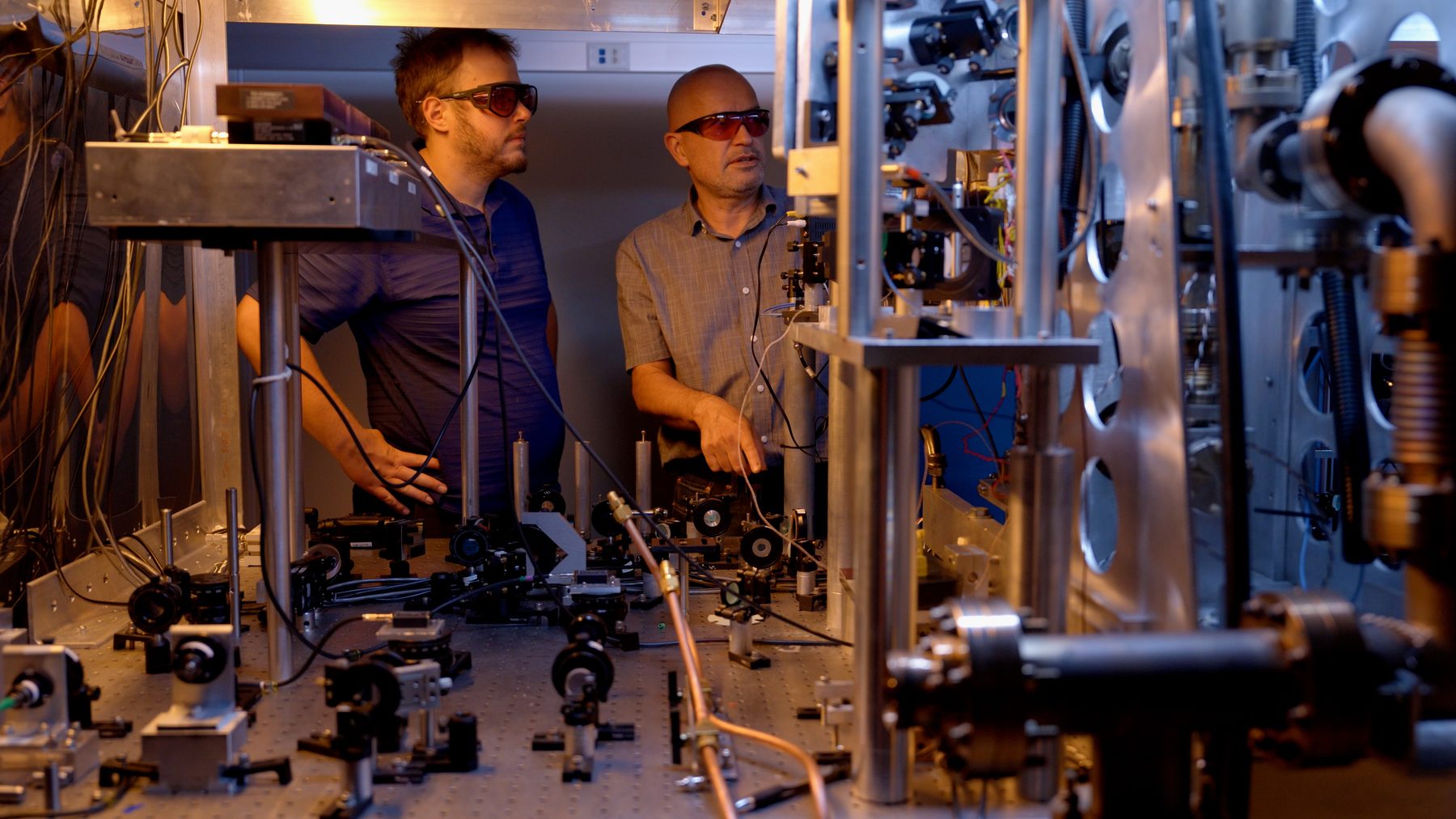 Two physicists in safety glasses at a bench of lasers and optics, part of the NIST-F4 caesium fountain clock.